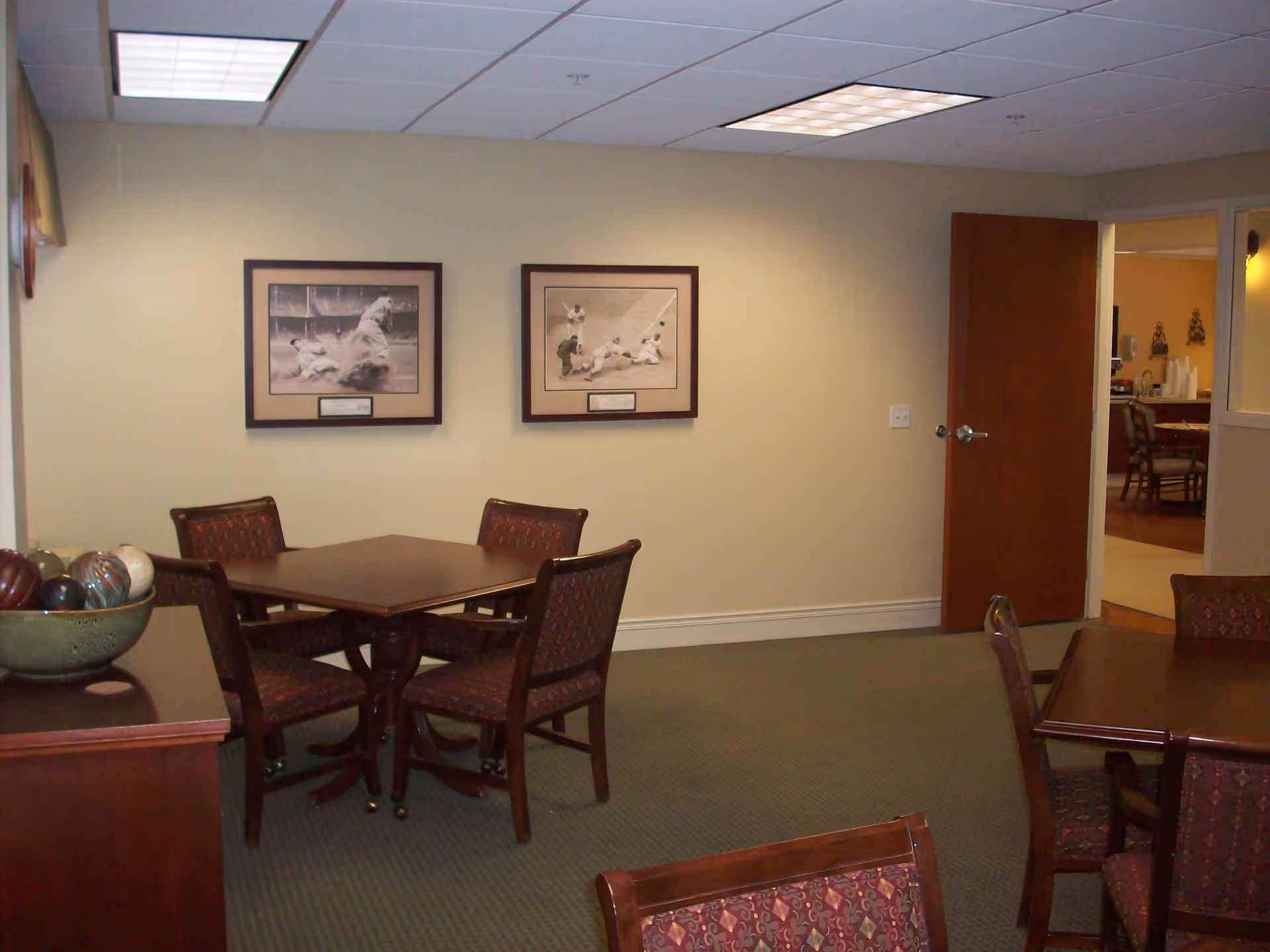 Interior room with two square wooden tables, each surrounded by four upholstered chairs with patterned fabric. Two framed black and white sports photographs hang on a beige wall. A wooden sideboard with a decorative bowl filled with ornamental balls is visible on the left. An open door leads to another room with more chairs and tables.