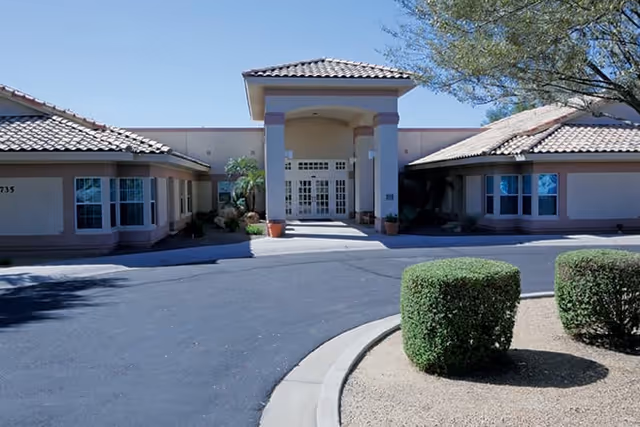 Front exterior view of a single-story building with a tiled roof and a covered entrance supported by columns. There are neatly trimmed bushes and desert landscaping in front of the building under a clear blue sky.