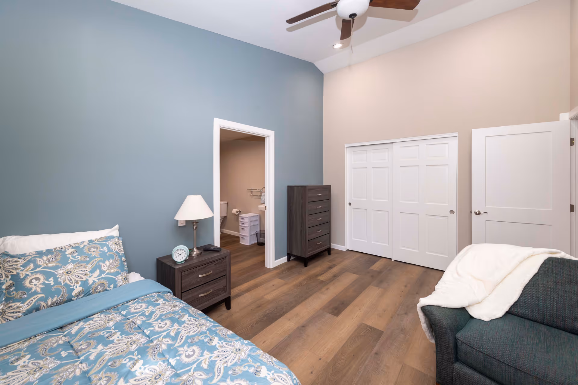 A bedroom with a bed covered in blue and white patterned bedding, a nightstand with a lamp and clock, a dresser, a green armchair with a white blanket draped over it, and an open door leading to a bathroom.