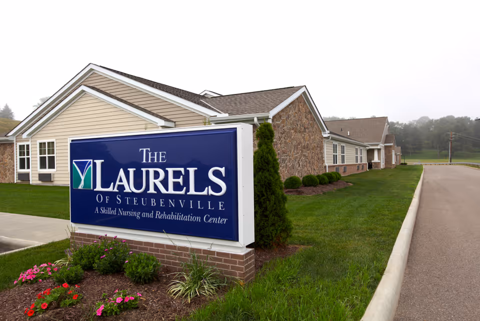 Exterior view of The Laurels of Steubenville nursing and rehabilitation center with a large blue sign in front of a single-story building and landscaped lawn.