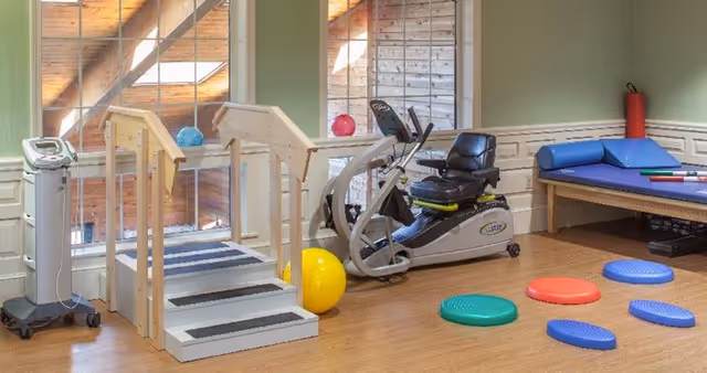 A senior living community exercise room with a small set of stairs, a recumbent exercise bike, colorful balance pads on the floor, a large yellow exercise ball, and a padded therapy table with blue cushions. The room has large windows letting in natural light and wooden flooring.
