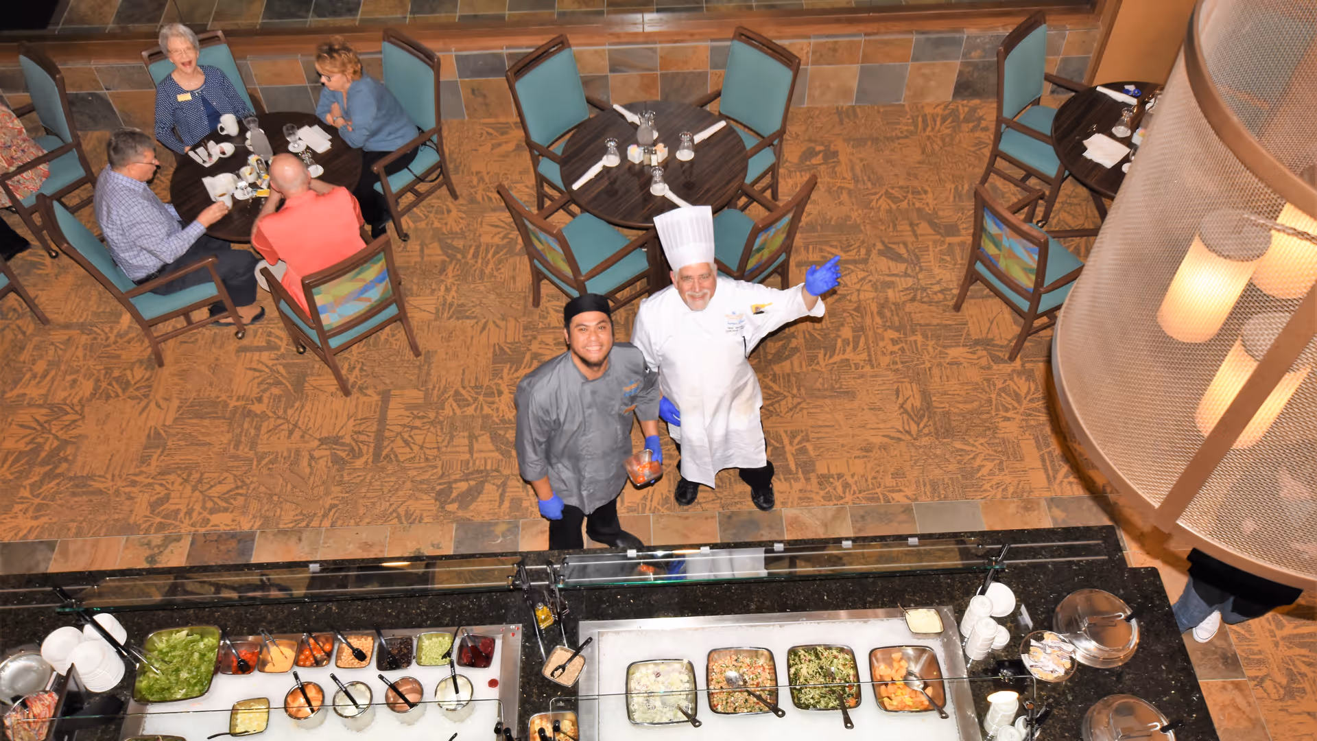 Overhead view of a dining area in a retirement resort with several tables and chairs. Two chefs, one in a white chef's coat and hat and the other in a gray chef's coat and black hat, are standing behind a buffet counter with various food items. Three seniors are seated at a table enjoying their meal and conversation.
