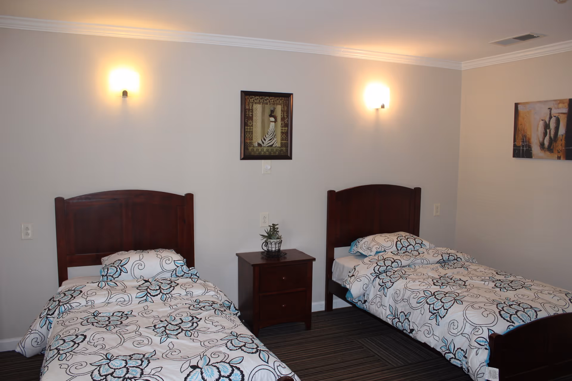 A bedroom with two single beds featuring dark wooden headboards and white bedspreads with blue and black floral patterns. A small wooden nightstand with two drawers and a small plant is placed between the beds. Two wall-mounted lights are above each bed, and two framed artworks hang on the walls. The room has light-colored walls and a striped carpeted floor.