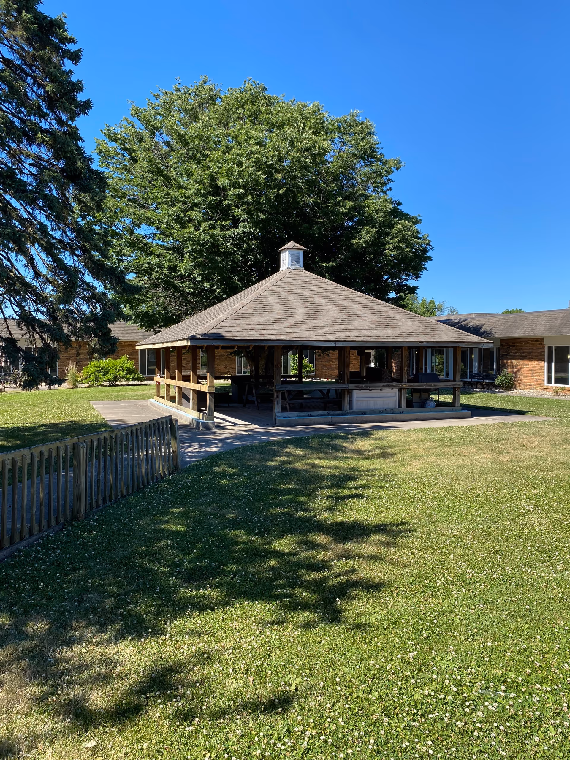 A wooden open-air pavilion with picnic tables sits in a grassy courtyard before a brick senior living building and large tree.