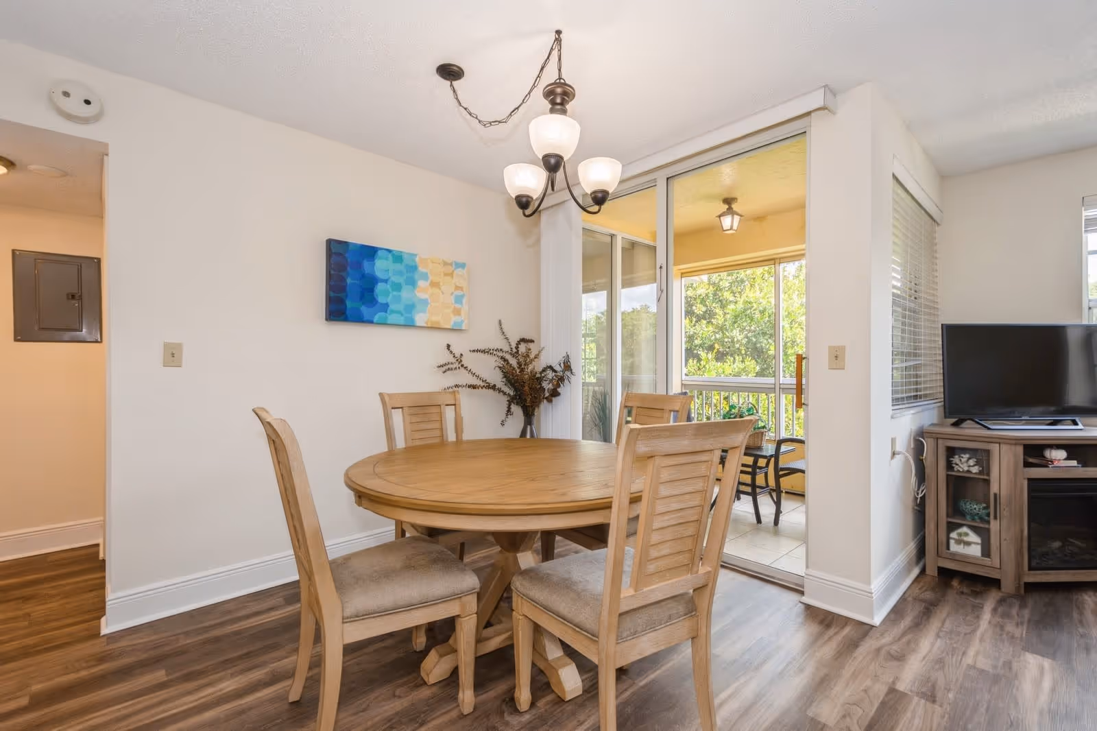 A dining area with a round wooden table and four matching chairs on a wood floor. A chandelier with three lights hangs above the table. There is a blue and beige abstract painting on the wall and a vase with dried plants on the table. Sliding glass doors lead to a small balcony with outdoor seating and greenery visible outside. To the right, part of a living room with a TV on a wooden stand is visible.