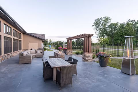 Patio with wicker dining table and chairs, lounge seating, a wooden pergola, planters, and a patio heater beside a building and fenced lawn.