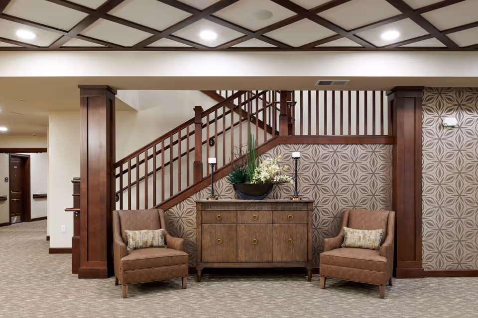 Seating area with two upholstered armchairs flanking a wooden console beneath a staircase in a warmly decorated common room.