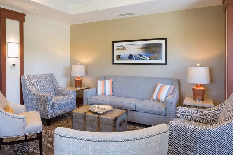 A cozy living room with a gray sofa adorned with two striped pillows, surrounded by four patterned armchairs. Two side tables with matching lamps flank the sofa. A framed picture of boats on water hangs on the beige wall above the sofa. The room has a patterned carpet and soft lighting.