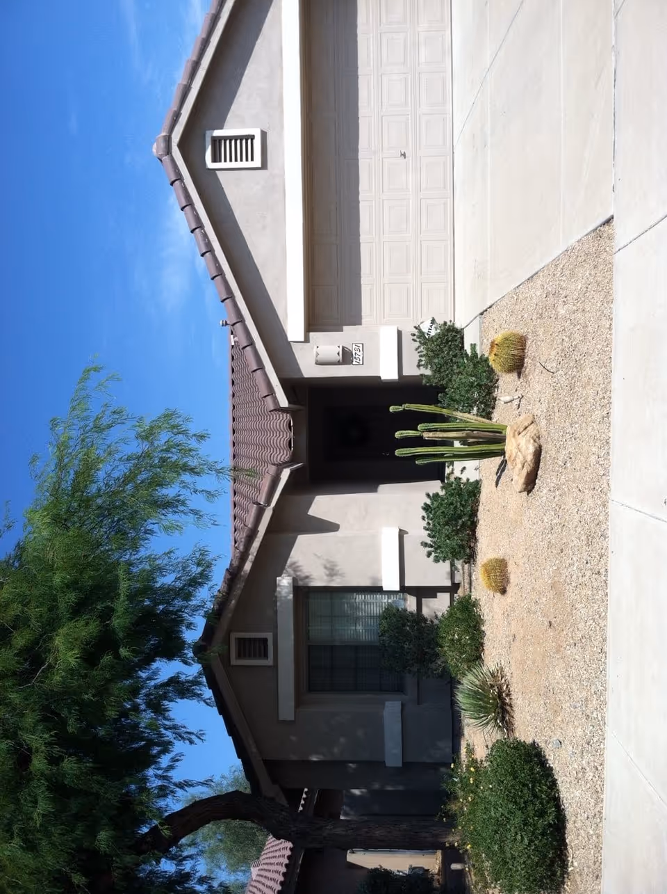Front exterior view of a single-story house with a beige stucco finish, a tiled roof, a garage door, and a small desert-style garden featuring cacti and shrubs. A tree is visible on the left side under a clear blue sky.