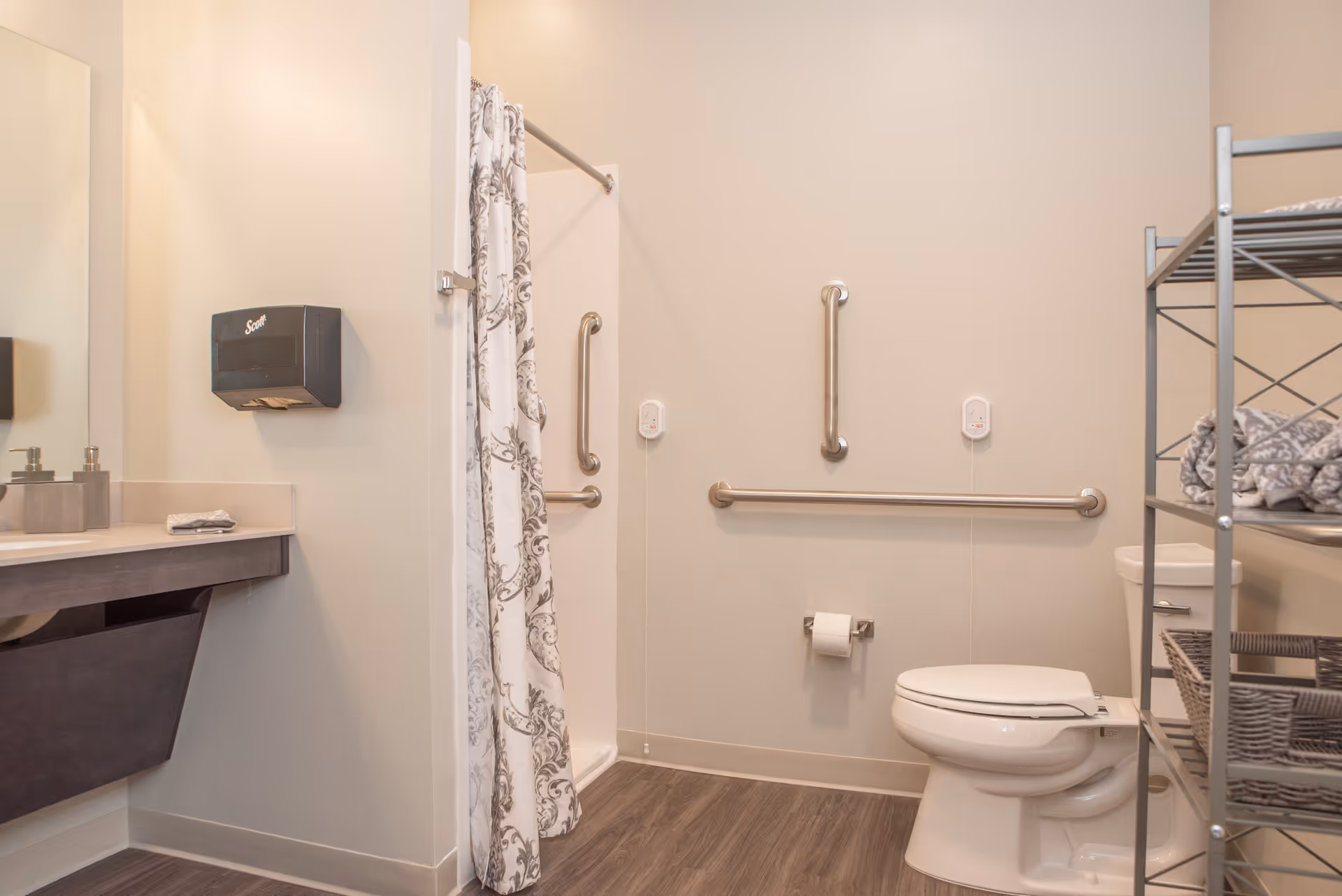 Accessible bathroom with a toilet, grab bars on the walls, a shower with a patterned curtain, a sink with soap dispensers, a paper towel dispenser, and a metal shelving unit holding folded towels and baskets.