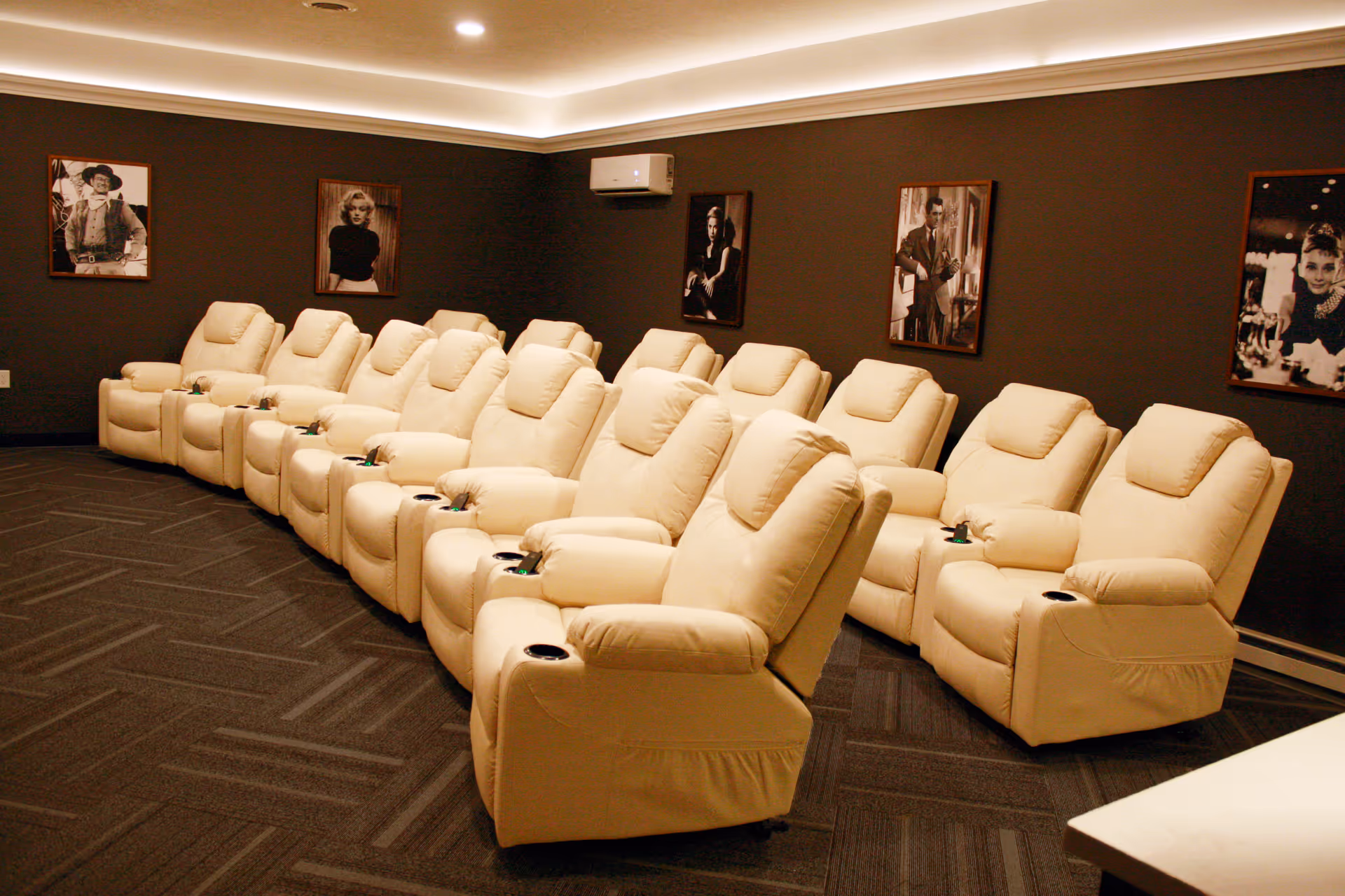 A cozy home theater room with two rows of cream-colored reclining chairs arranged facing forward. The walls are dark with framed black and white portraits of classic movie stars. The ceiling has recessed lighting with a soft glow.