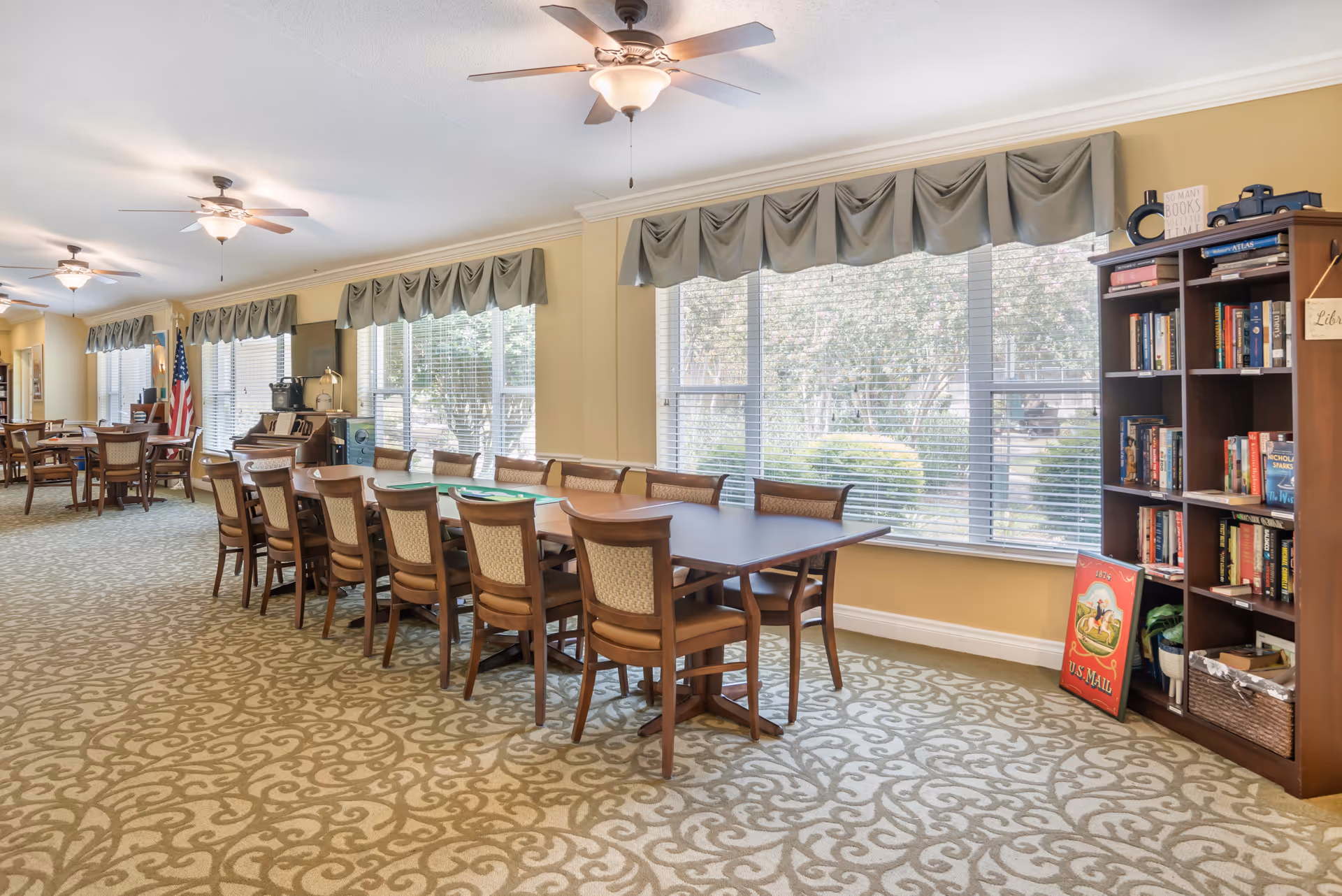 A spacious room with large windows covered by valance curtains, featuring a long table with multiple wooden chairs arranged around it. The room has patterned carpet flooring, ceiling fans with lights, and a bookshelf filled with books and decorative items. An American flag is visible in the background near another set of tables and chairs.