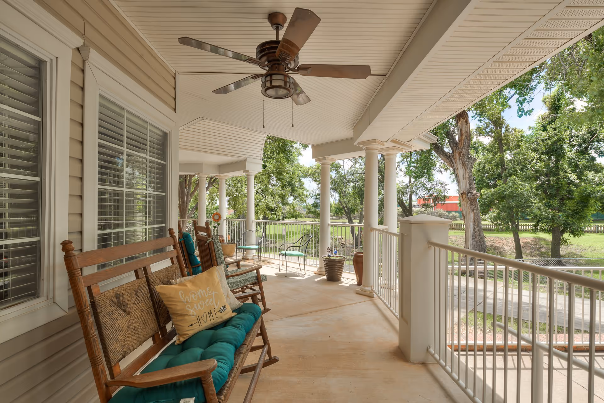 A covered outdoor porch area with wooden rocking chairs and a bench with cushions, including a pillow that says 'home sweet home'. The porch has white columns, a ceiling fan, and overlooks a green yard with trees and a pathway.