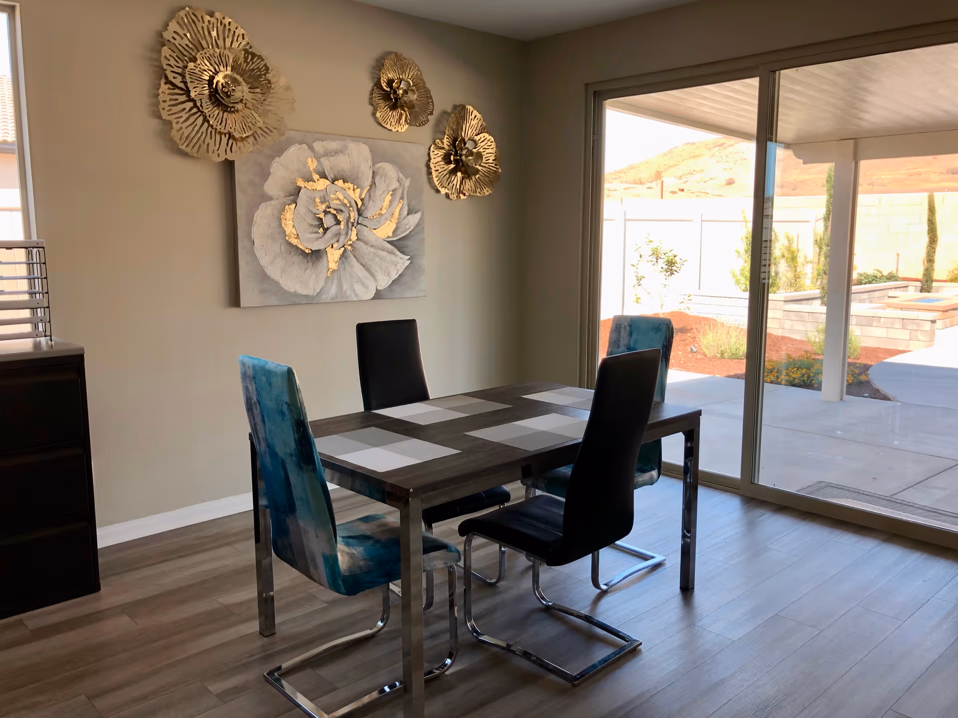 A modern dining room with a rectangular table set for four, featuring two black chairs and two blue patterned chairs. The wall has decorative gold and gray floral artwork. Large sliding glass doors open to an outdoor patio with a view of a garden and hills in the background.