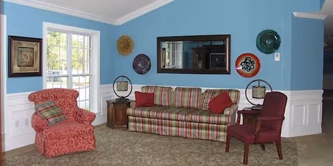 A cozy living room with light blue walls and white wainscoting. The room features a striped sofa with red and green cushions, a red patterned armchair, and a red wooden chair. There are decorative plates and a mirror hanging on the wall, and a window letting in natural light.