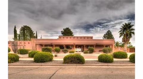 Exterior view of a single-story building with a flat roof and adobe-style architecture, surrounded by neatly trimmed bushes and trees under a cloudy sky.