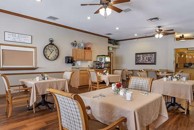 A dining room with several tables covered in beige tablecloths, each set with cups, utensils, and small flower arrangements. The room has wooden chairs with cushioned seats and backs, ceiling fans with lights, a wall clock, and a beverage station with cabinets and a coffee machine. The walls are decorated with framed artwork and a sign that reads 'BLESS the food before us, the family beside us, and the love between us.'