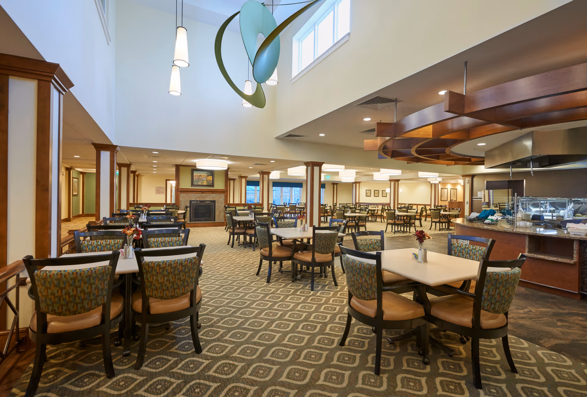 Spacious dining room in a retirement community with multiple tables and chairs arranged neatly. The room features a patterned carpet, a fireplace, large windows near the ceiling allowing natural light, and a serving area with a counter and food service equipment. Modern pendant lights hang from the ceiling.
