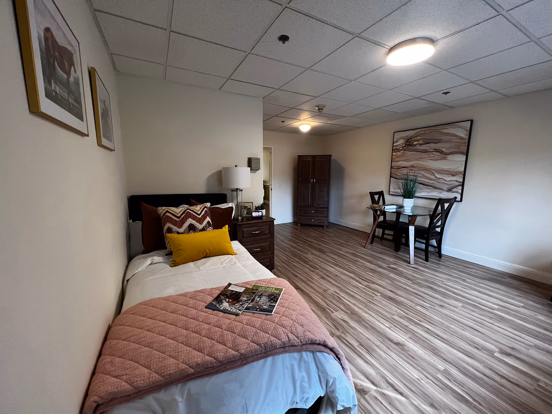 Furnished memory-care studio room with a single bed, nightstand and lamp, armoire, and a small dining table on wood-look flooring.