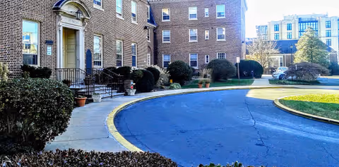 Curved driveway leading to the entrance of a multi-story brick building with several windows. There are bushes and potted plants along the sidewalk near the entrance. Another building is visible in the background under a clear sky.