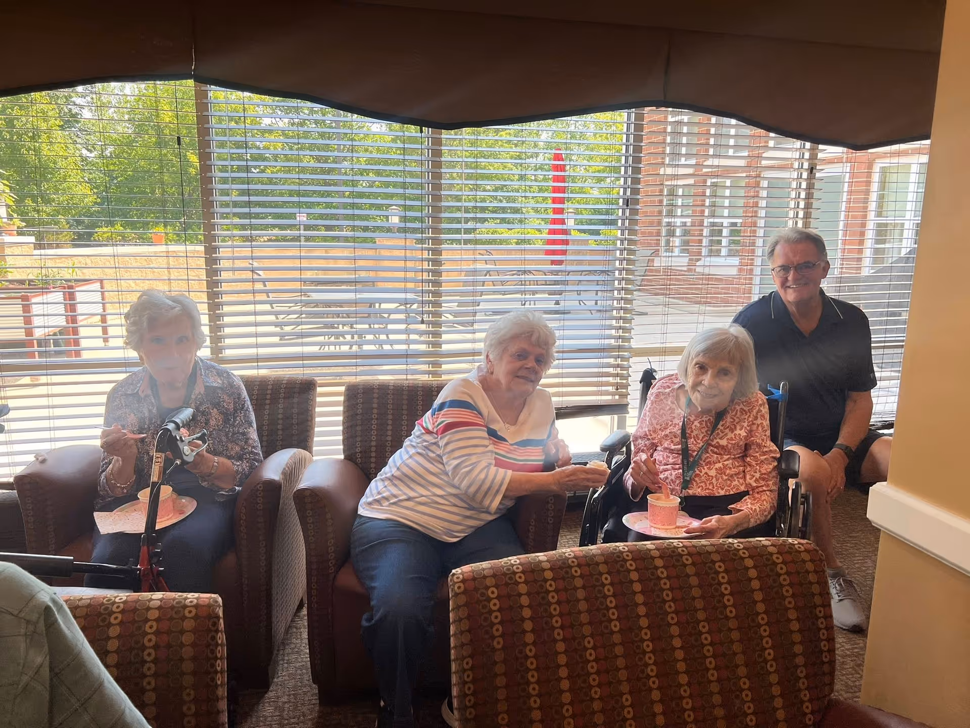 Four elderly individuals sitting in a living room area with large windows and blinds behind them. Two women are seated in armchairs, one woman in a wheelchair, and a man is sitting on a chair. They appear to be enjoying ice cream or dessert together. Outside the window, there is a patio with tables, chairs, and a red umbrella.