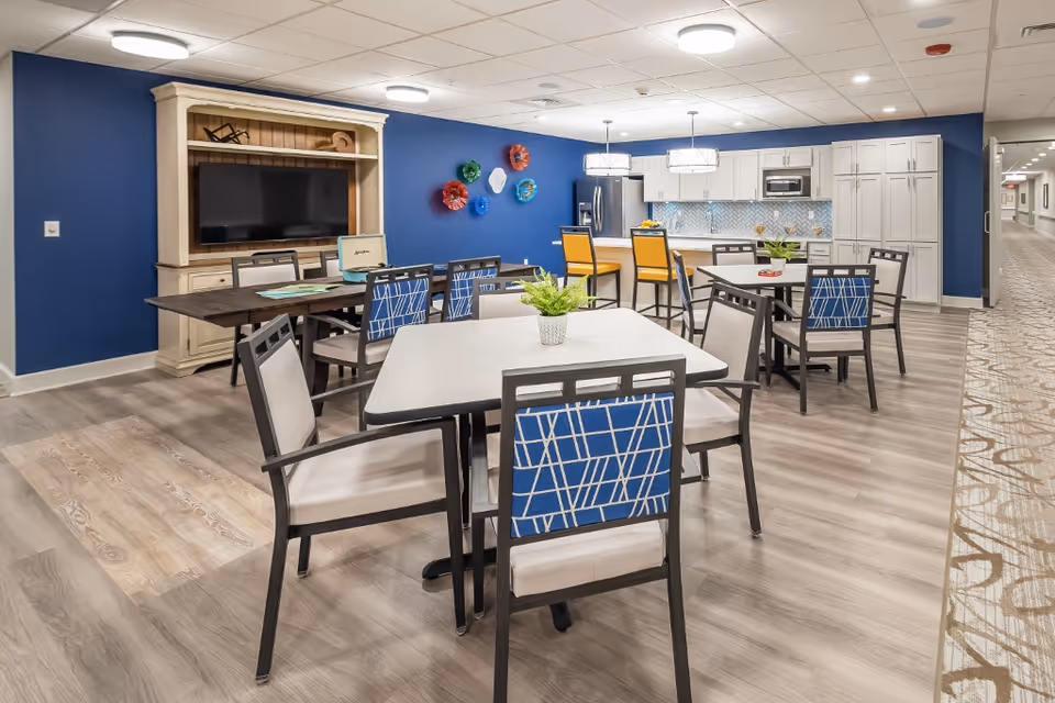 A bright and modern common area in a senior living facility featuring multiple tables and chairs with blue and beige upholstery. The room has a blue accent wall with colorful decorative plates, a large TV mounted on a cream-colored entertainment unit, and a kitchen area with white cabinets, a refrigerator, microwave, and a breakfast bar with yellow chairs. The flooring is light wood laminate, and the ceiling has recessed lighting.