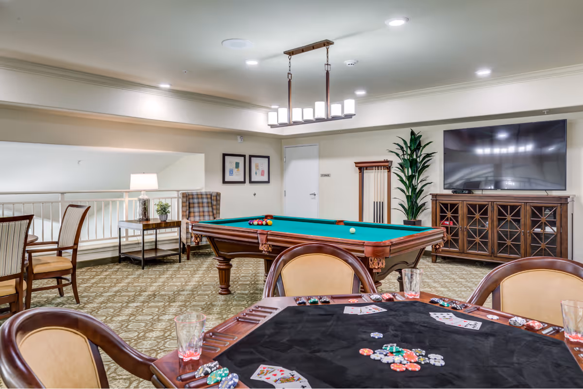 A recreational room featuring a pool table with billiard balls arranged for a game, a poker table with playing cards and poker chips, several chairs, a large flat-screen TV mounted on a wooden cabinet, a tall green plant, and a side table with a lamp and small plant. The room has carpeted flooring and recessed ceiling lights.