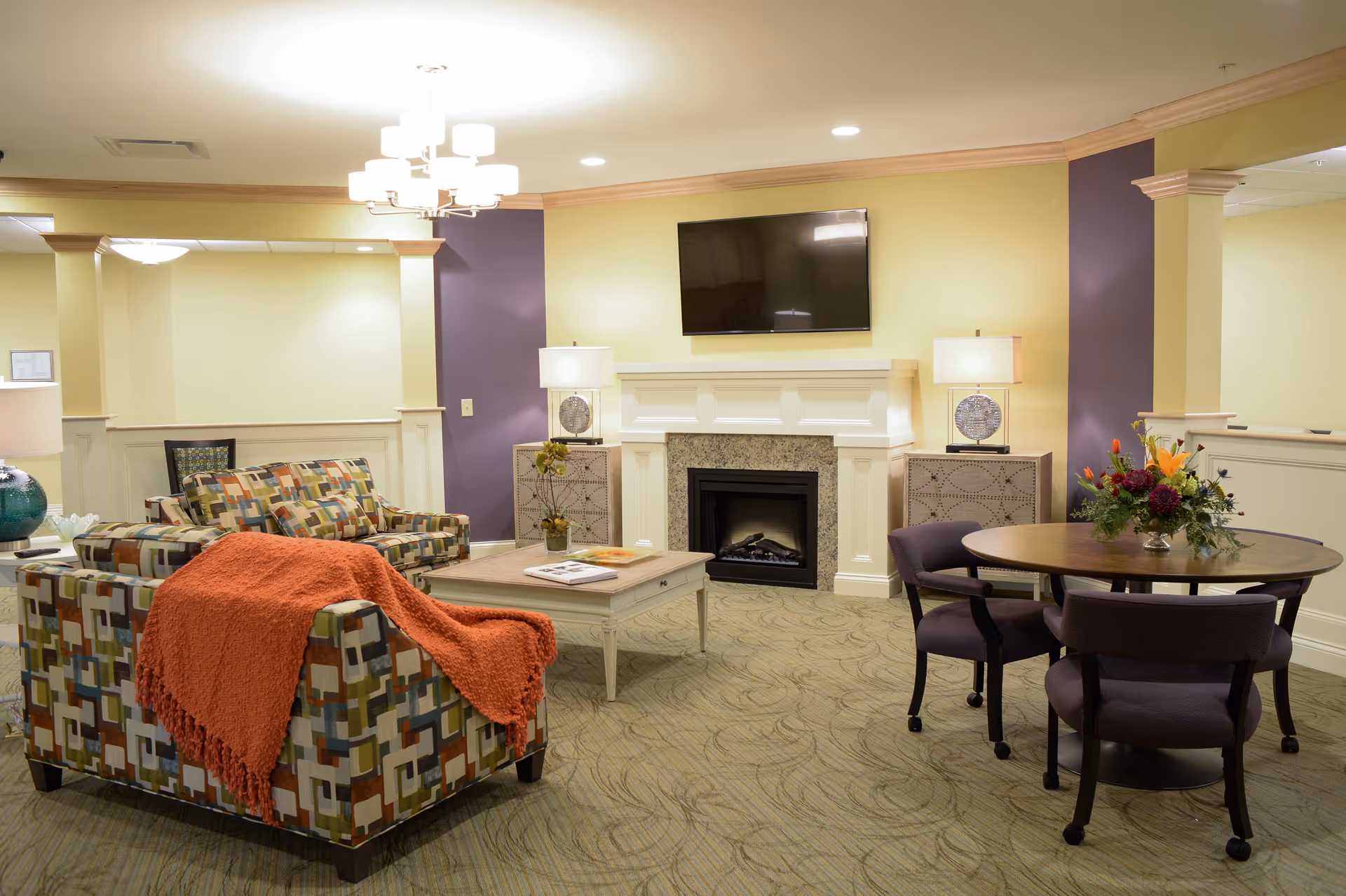 Cozy communal living room with patterned sofas, a coffee table, fireplace and wall-mounted TV, and a small round table with chairs.