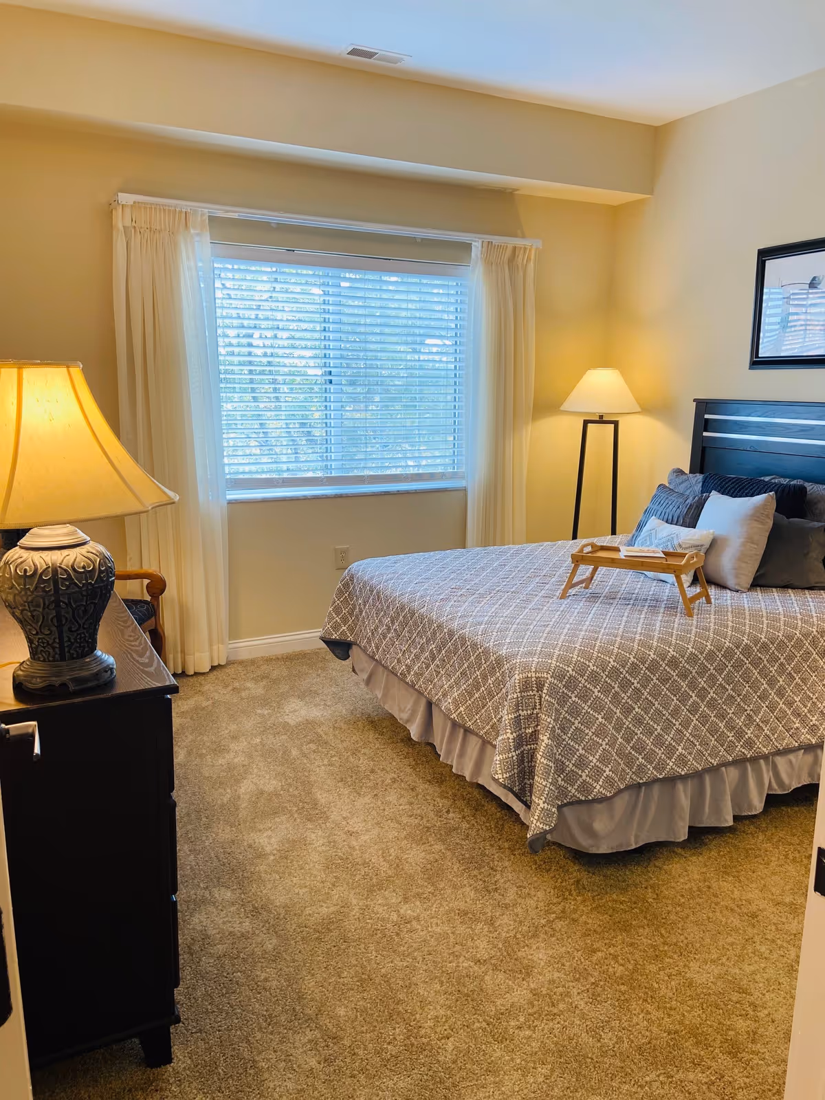 A cozy bedroom with a large window covered by white curtains and blinds. The room features a bed with a patterned bedspread and multiple pillows, a wooden tray on the bed, a dark wooden dresser with a decorative lamp, and a floor lamp in the corner. The walls are painted a light beige color and the floor is carpeted.