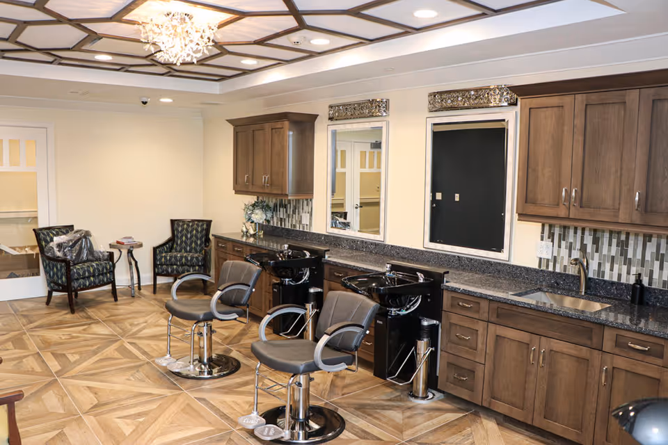 Interior view of a salon area in a senior living facility featuring two black salon chairs in front of two black wash basins with mirrors above. The room has wooden cabinets, a granite countertop with a sink, patterned tile backsplash, and a decorative ceiling with a chandelier. There are two upholstered chairs and a small table in the corner.