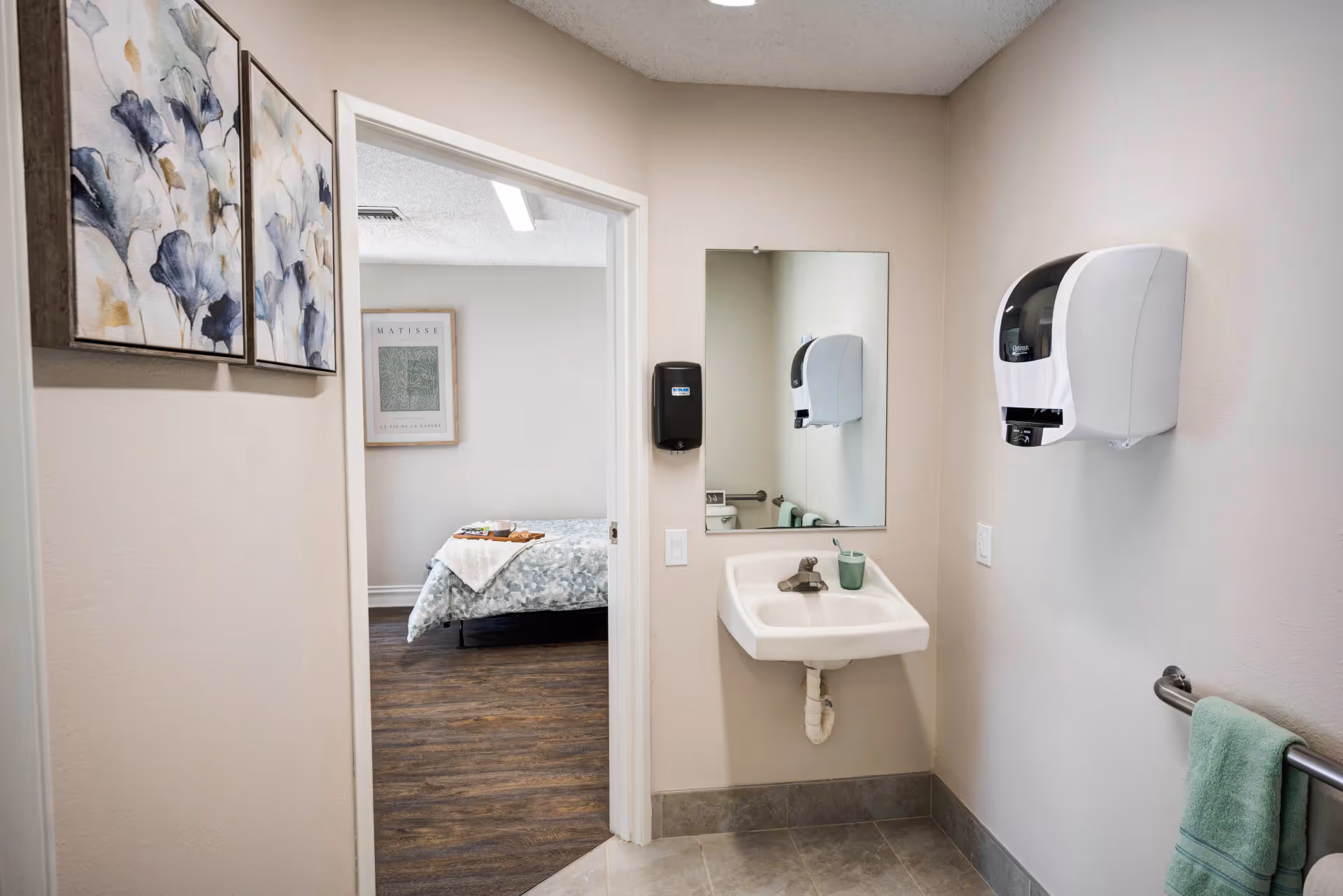 A small bathroom area with a wall-mounted sink, a mirror above it, a soap dispenser, and a towel rack with a green towel. The bathroom opens into a bedroom with a bed covered in a blue and white patterned blanket and a tray with a cup and plate on it. The walls are light-colored, and there are two framed floral paintings on the left wall near the doorway.