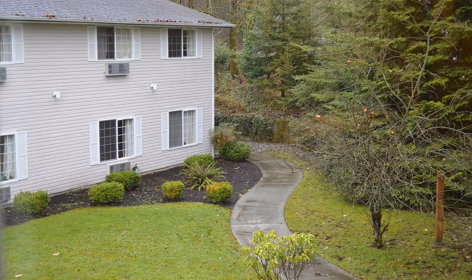 A side view of a two-story beige building with multiple windows and air conditioning units. In front of the building is a landscaped area with bushes and a curved concrete pathway leading into a wooded area with trees and grass.