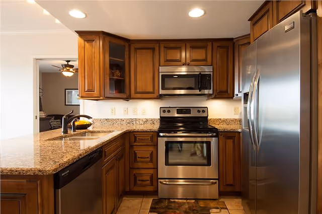 A modern kitchen with wooden cabinets, granite countertops, a stainless steel refrigerator, oven, microwave, and a dishwasher. The kitchen has recessed lighting and a sink with a faucet on the left side. An adjoining room with a ceiling fan and framed pictures is visible through an open doorway.