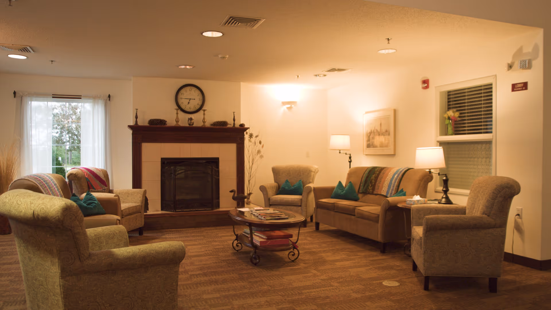 A cozy living room with a fireplace in the center, surrounded by several upholstered armchairs and a sofa with colorful throw blankets and teal pillows. A round coffee table with magazines sits in the middle of the seating area. The room is softly lit by table lamps and a wall sconce, with windows on two walls allowing natural light.
