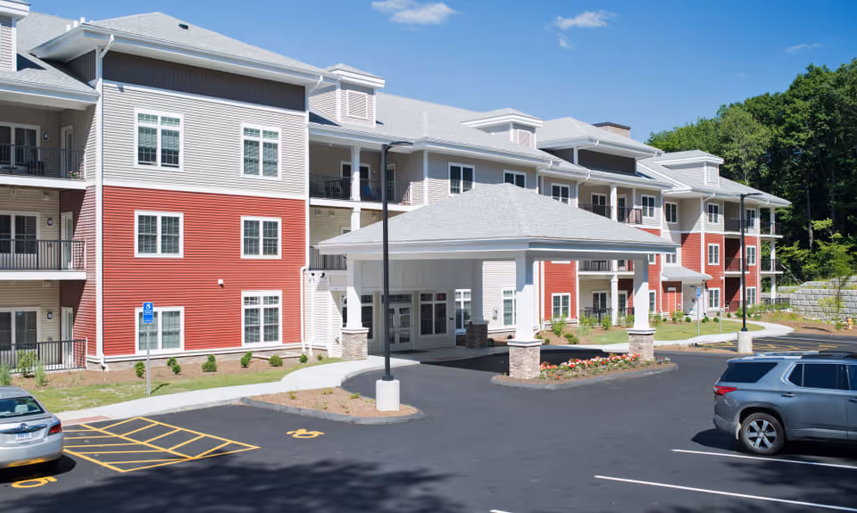 Exterior view of a multi-story senior living facility building with red and beige siding, multiple windows, balconies, a covered entrance, and a parking lot with several cars parked. Trees and greenery are visible in the background under a clear blue sky.