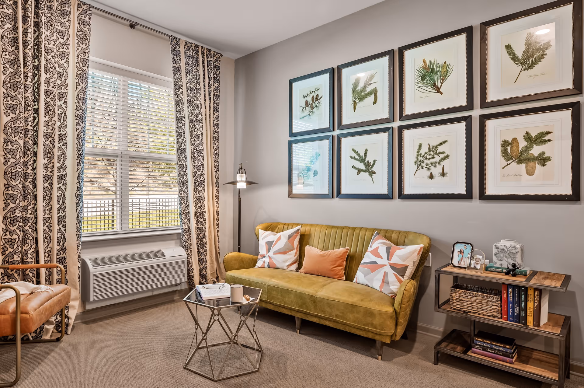 A cozy living room with a mustard yellow sofa adorned with three decorative pillows. Above the sofa are eight framed botanical prints arranged in two rows. To the right of the sofa is a small wooden and metal bookshelf holding books, a basket, and framed photos. To the left is a window with patterned curtains and a wall-mounted air conditioning unit below it. A modern floor lamp stands next to the sofa, and a small geometric glass-top coffee table is in front of the sofa.