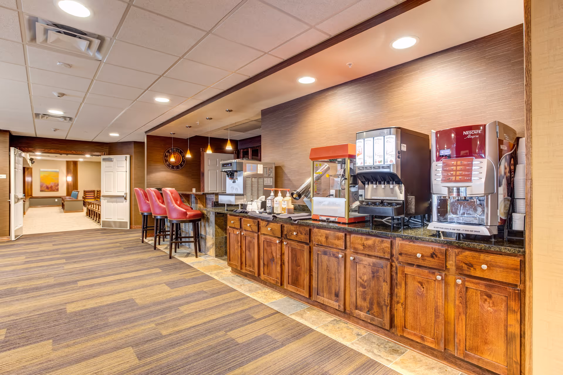 Interior view of a beverage and snack counter area in a senior living facility with wooden cabinets, a popcorn machine, a soda dispenser, a coffee machine, and three red bar stools. The area is well-lit with ceiling lights and pendant lights, and there is an open doorway leading to another room with seating and artwork on the walls.