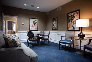 A cozy interior room with a gray sofa on the left, a small round table with four chairs in the center, and two wooden chairs with a small round side table and a lamp on the right. The walls are beige with white wainscoting, and there are framed vintage-style posters hanging on the walls.