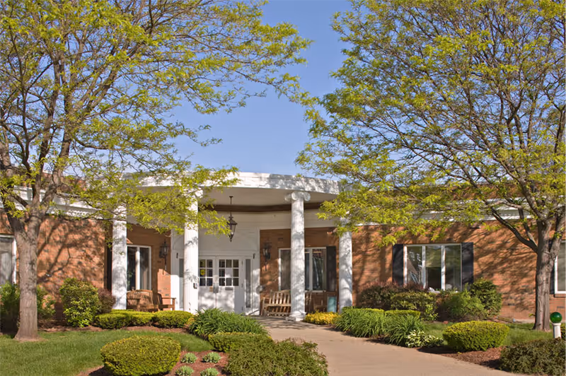 Front entrance of a single-story brick building with white columns and a covered porch. There are benches on either side of the entrance, surrounded by well-maintained bushes and trees with green leaves under a clear blue sky.