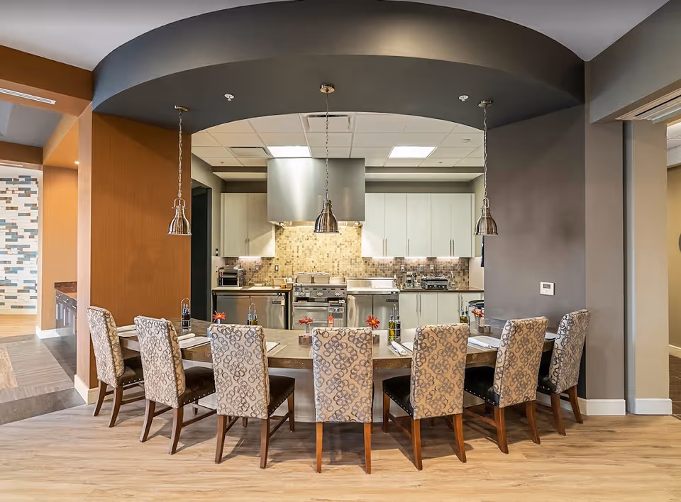 A modern kitchen area with a long dining counter and eight upholstered chairs arranged in front. The kitchen features stainless steel appliances, white cabinets, and a tiled backsplash. Three pendant lights hang from a curved ceiling above the counter. The flooring is light wood, and the walls are painted in warm neutral tones.