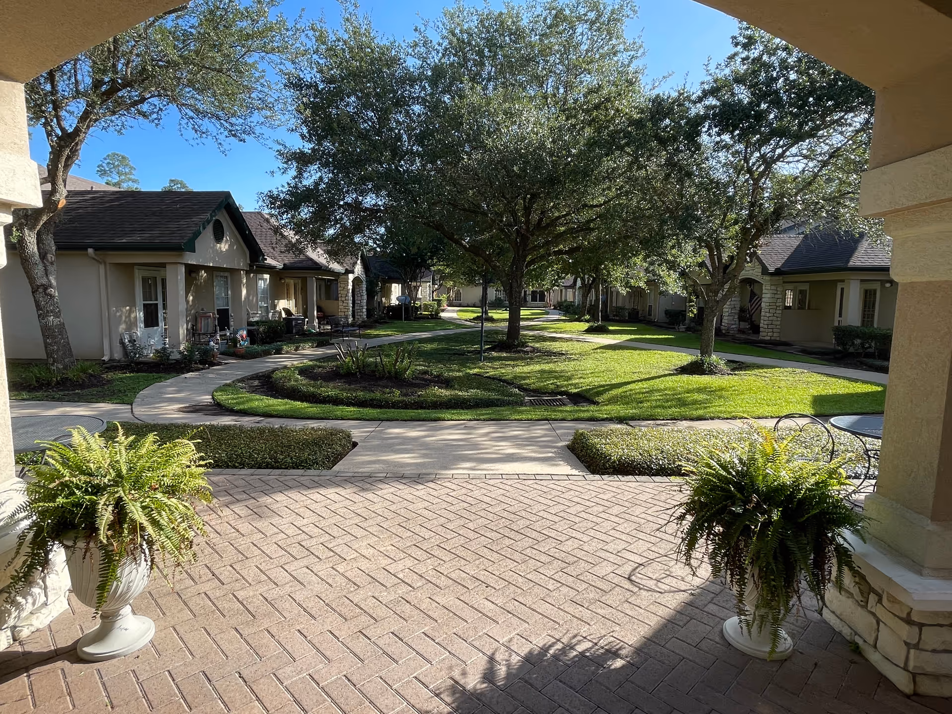 View from a covered patio area looking out onto a landscaped courtyard with green grass, trees, and paved walkways. Single-story residential buildings with porches surround the courtyard. Two large potted ferns are placed on either side of the patio entrance.