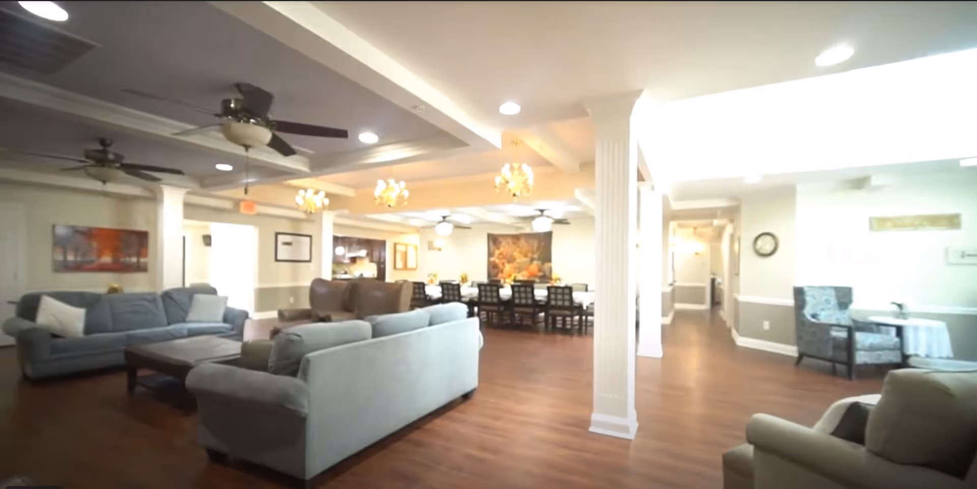 Spacious common area in a senior living facility featuring multiple seating arrangements including sofas and armchairs, ceiling fans, chandeliers, and a dining area with a long table and chairs. The room has wooden flooring, light-colored walls, and decorative artwork on the walls.