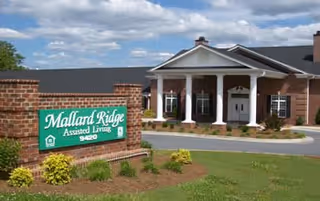 Exterior view of Mallard Ridge Assisted Living facility showing a brick building with white columns at the entrance and a green sign in front with the facility name and address.
