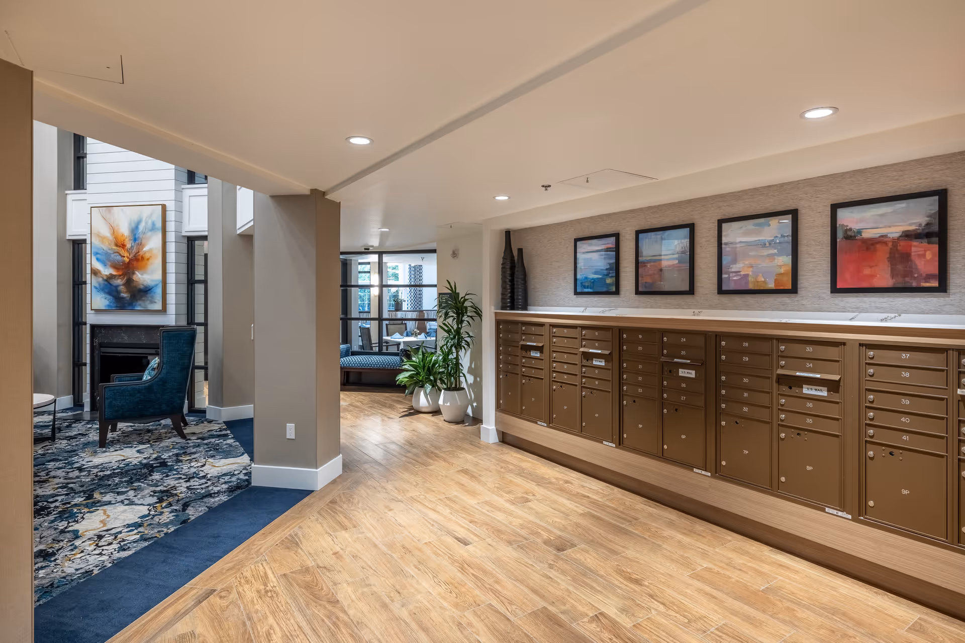 Interior view of a senior living facility hallway featuring a row of mailboxes on the right wall with four colorful abstract paintings above them. The floor is wood with a transition to carpeted seating area on the left, which includes a blue armchair and a fireplace with a large abstract painting above it. There are also some potted plants and recessed ceiling lights.