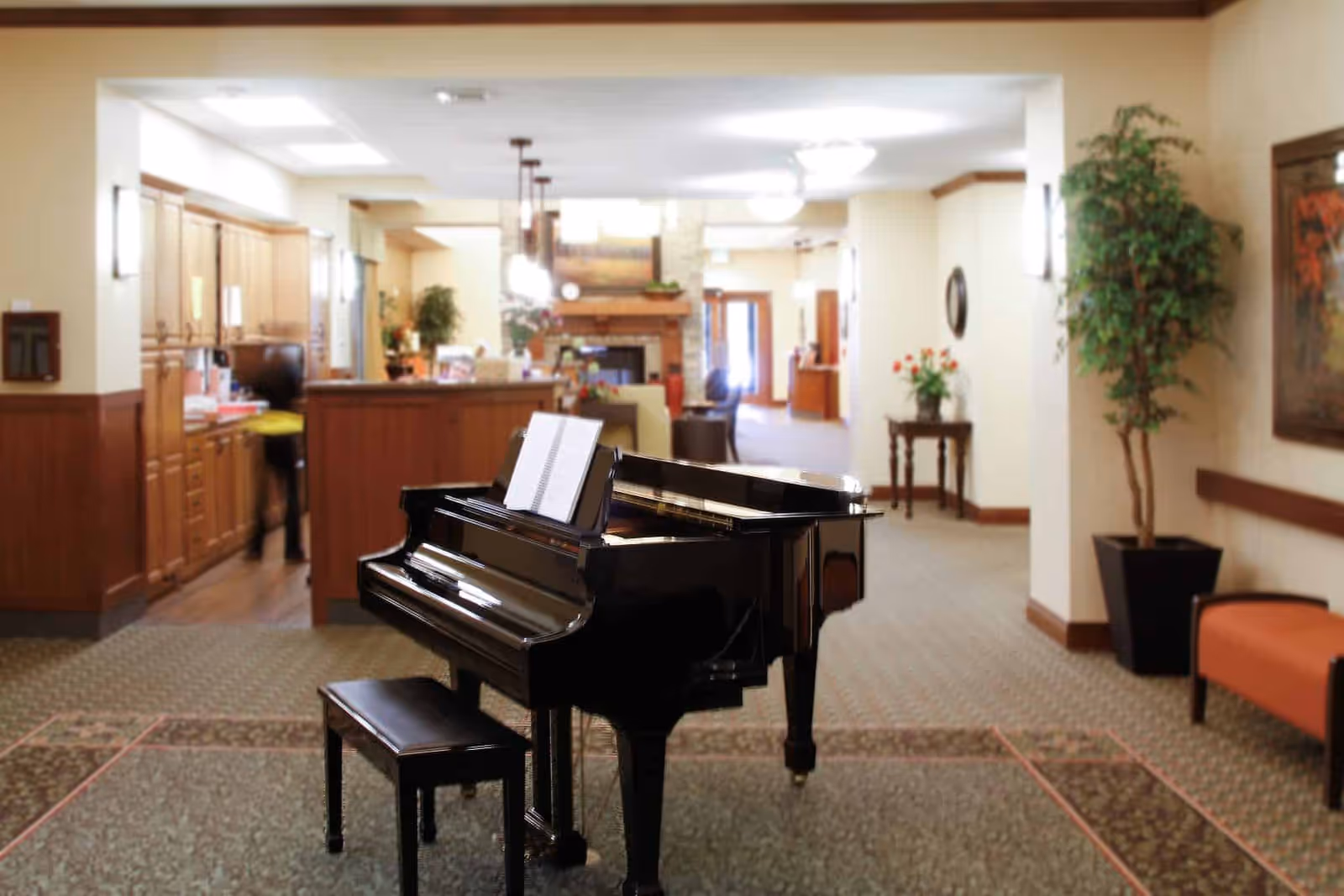 A black grand piano with an open music book stands in a senior living facility lobby with a reception desk, seating, and potted plants in the background.
