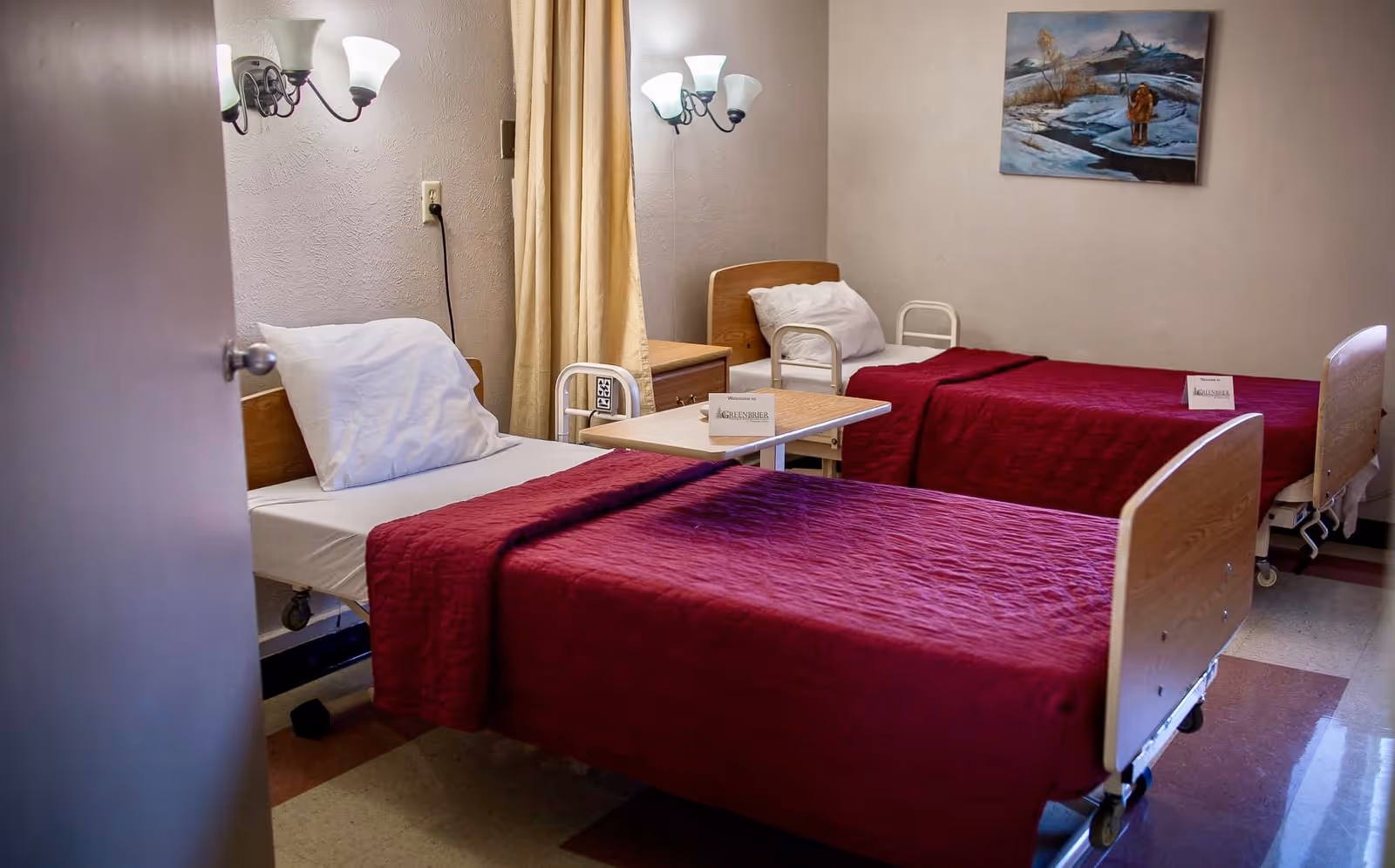 A nursing home patient room with two hospital-style beds made with red bedspreads, bedside tables, wall-mounted lamps, and a painting on the wall.