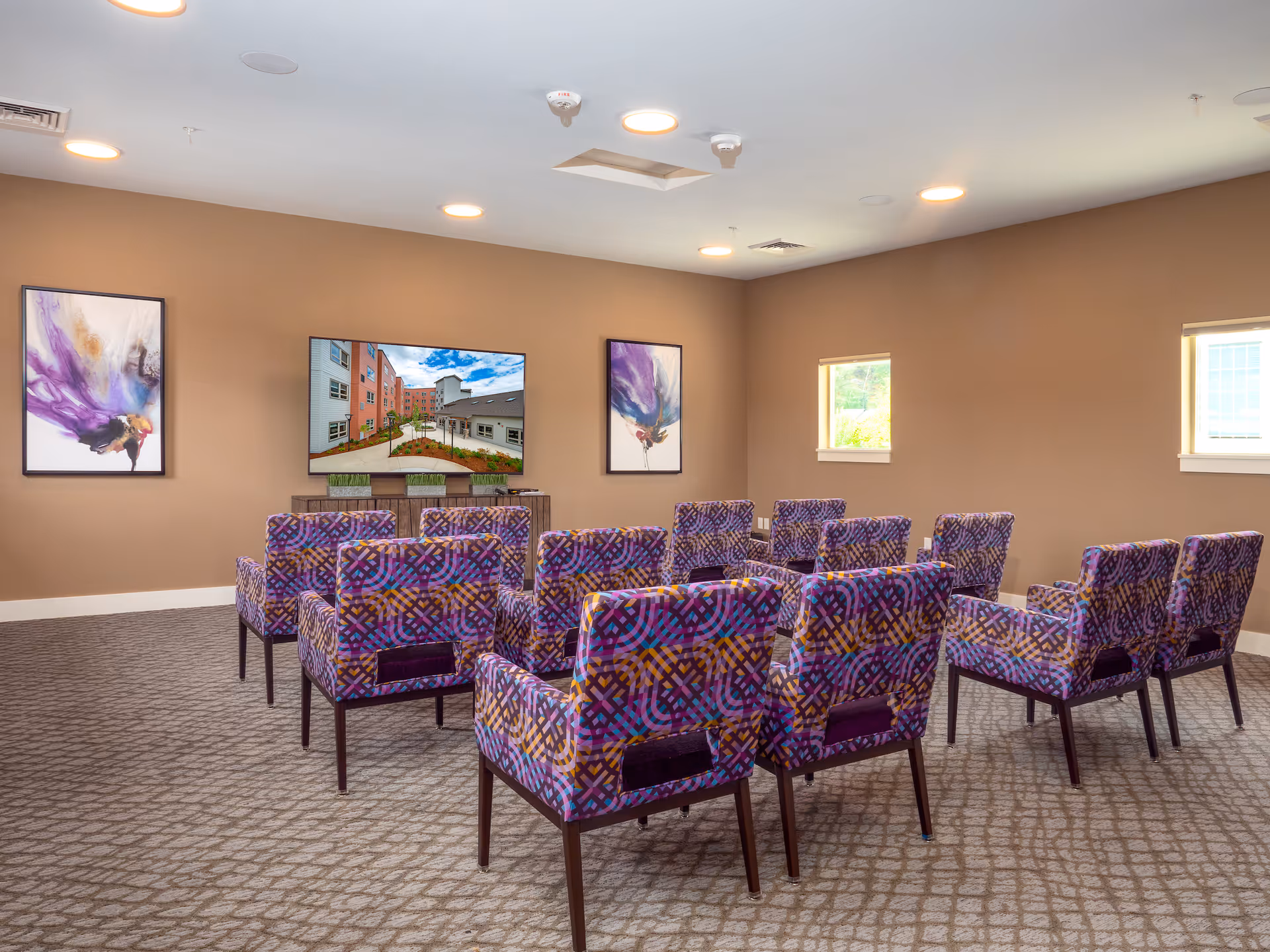 A small theater or media room with three rows of colorful patterned chairs facing a large flat-screen TV mounted on a beige wall. The room has carpeted flooring, two small windows on the right wall, and two abstract paintings on the front wall beside the TV.