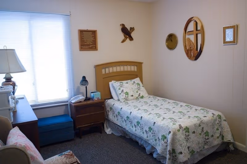 A small, cozy bedroom with a single bed covered in a floral quilt. The room has a wooden headboard, a nightstand with a lamp and a telephone, a window with blinds, and various wall decorations including a wooden eagle, plaques, and an oval mirror with a cross design.