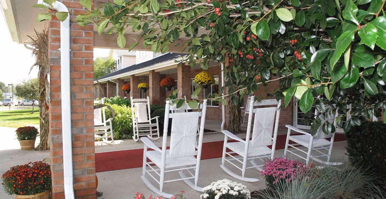 Covered outdoor patio area with white rocking chairs arranged on a concrete floor. The patio is decorated with hanging flower baskets and potted flowers, surrounded by green bushes and trees. A brick column supports the roof of the patio.