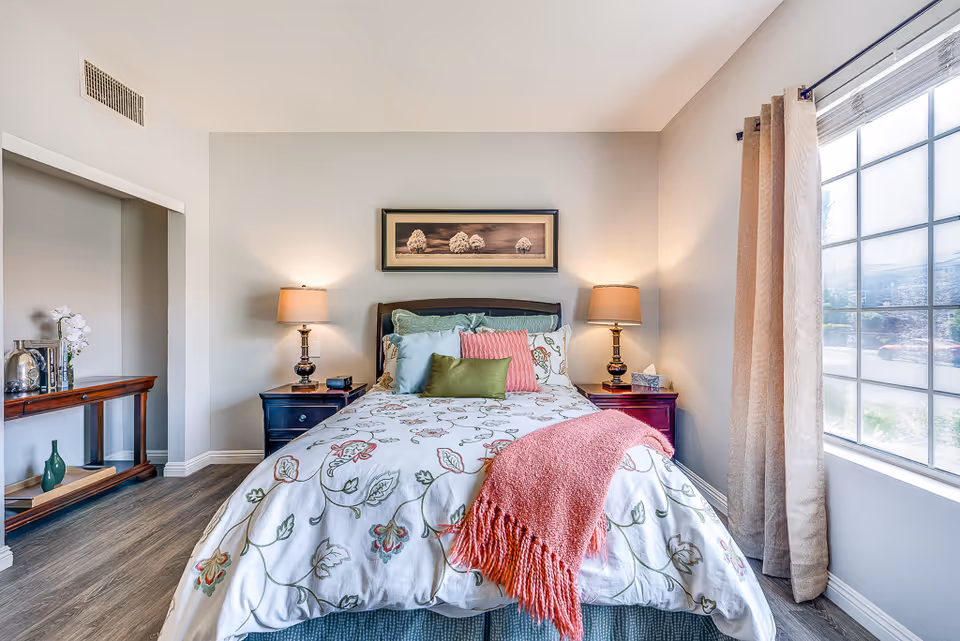 A bright and cozy bedroom with a bed centered against a light gray wall. The bed has a floral patterned white comforter, multiple pillows in green, pink, and white, and a coral pink throw blanket. There are two matching dark wood nightstands on either side of the bed, each with a lamp. A framed picture of trees hangs above the bed. To the right, a large window with beige curtains lets in natural light. To the left, a wooden console table with decorative items is visible.
